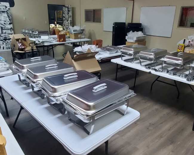 A spacious room is filled with several tables set up with chafing dishes, food containers, and scattered supplies.