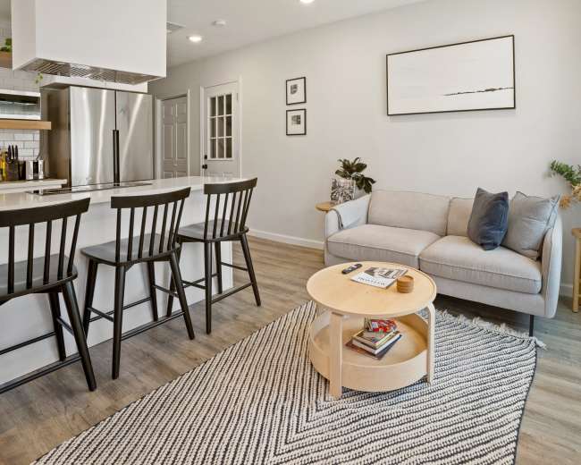 The image shows a modern living space featuring a light gray sofa, a round wooden coffee table with books, a woven rug, and a kitchen bar with three black stools.