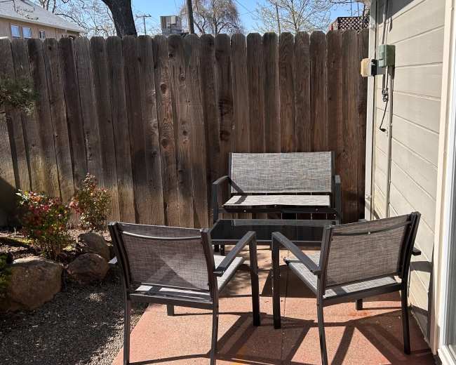 A small patio area features two chairs and a loveseat positioned against a wooden fence, surrounded by gravel and some greenery.