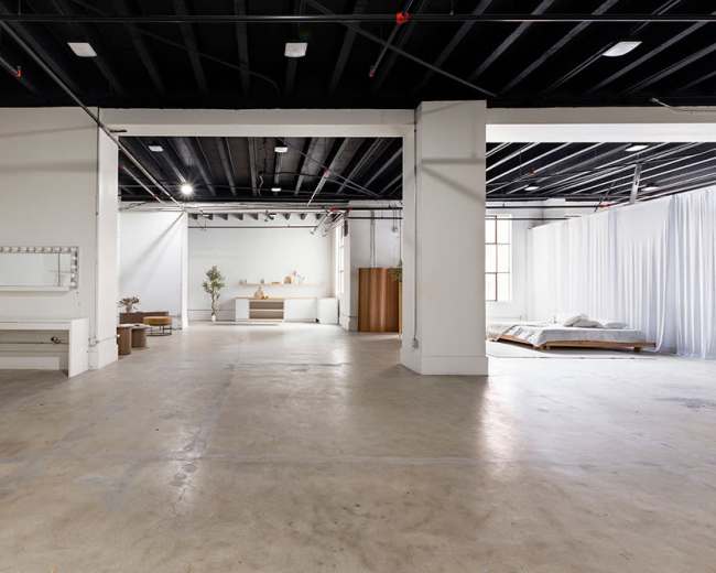 The image shows a spacious, empty interior with polished concrete flooring, white walls, and a minimalist design featuring a bed, a dressing table with lights, and draped fabric partitions.