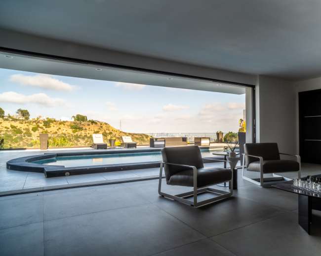 A modern living room opens to a pool area with a scenic view of hills and lounge chairs.