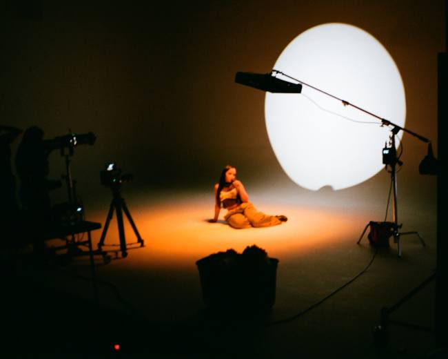A person sits on the floor of a studio, illuminated by a large circular light in the background.