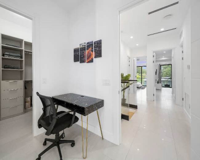 The image shows a modern hallway with a desk and chair alongside a closet on one side and a view of a bright living area at the end.