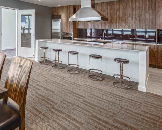 The image shows a modern kitchen and dining area featuring a long wooden table, high-backed chairs, a sleek kitchen island with stools, and large windows letting in natural light.