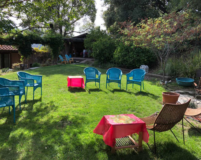 A backyard space features several blue chairs arranged in a circle around pink tables on green grass, with trees and a building in the background.