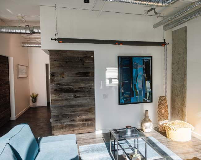 The image shows a modern interior space featuring a blue sofa, a glass coffee table, and a large framed artwork on the wall, with exposed ductwork and wooden accents.