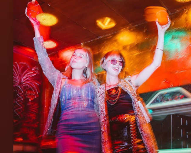 Two women are celebrating in a vibrant nightclub, holding drinks and wearing sequined outfits.