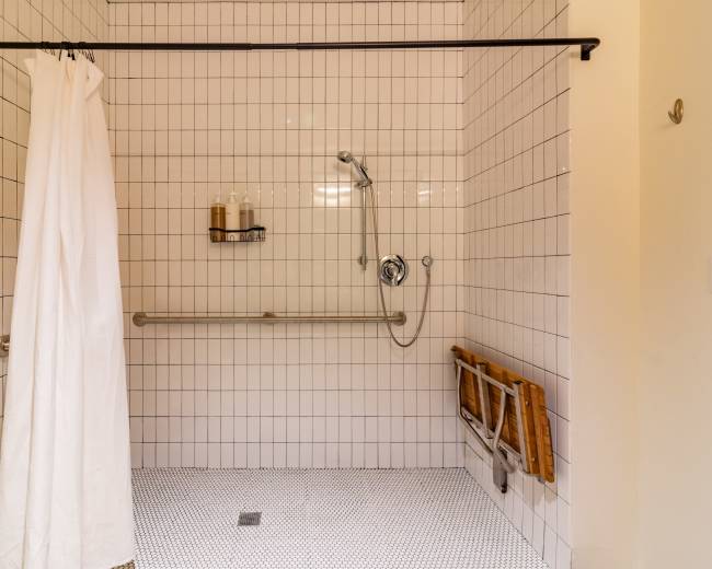 The image shows a tiled shower space with a handheld showerhead and a wall shelf for toiletries, featuring a white curtain and a folding bench made of wood.
