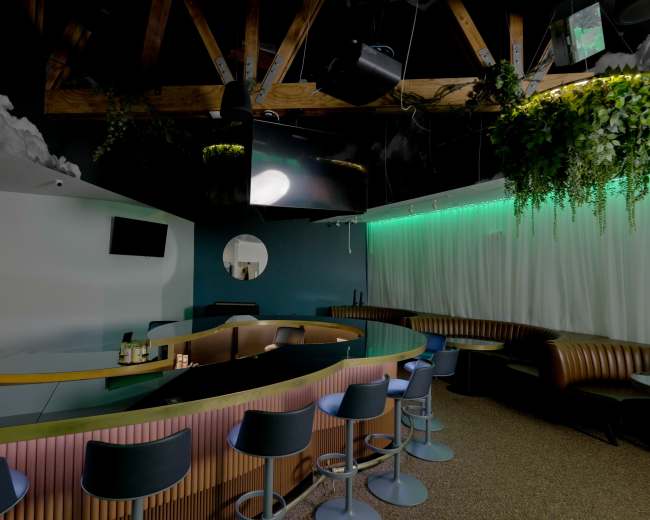 The image shows a modern bar interior featuring a curved counter, high stools, and a backdrop with green ambient lighting and lush greenery.