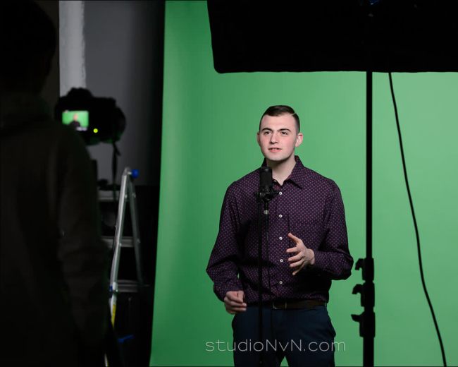 A person stands in front of a green screen, speaking into a microphone while being filmed.