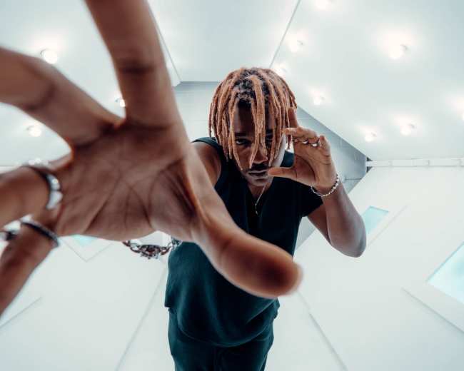 A person with dreadlocks holds a phone to their ear while extending one hand toward the camera in a white, brightly lit space.