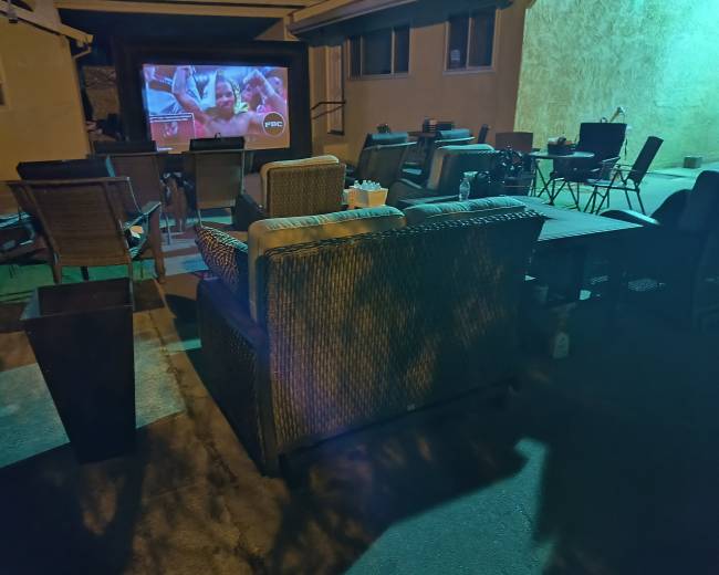The image shows an outdoor seating area with chairs and a couch facing a large screen displaying a movie or program against a wall.