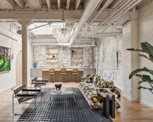 The image shows a modern living space with a patterned couch, stylish chairs, a bar area, and exposed brick walls.