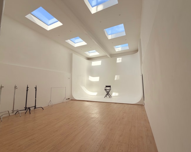 The image shows a spacious photography studio with a curved white backdrop, wooden flooring, and several skylights allowing natural light to enter.