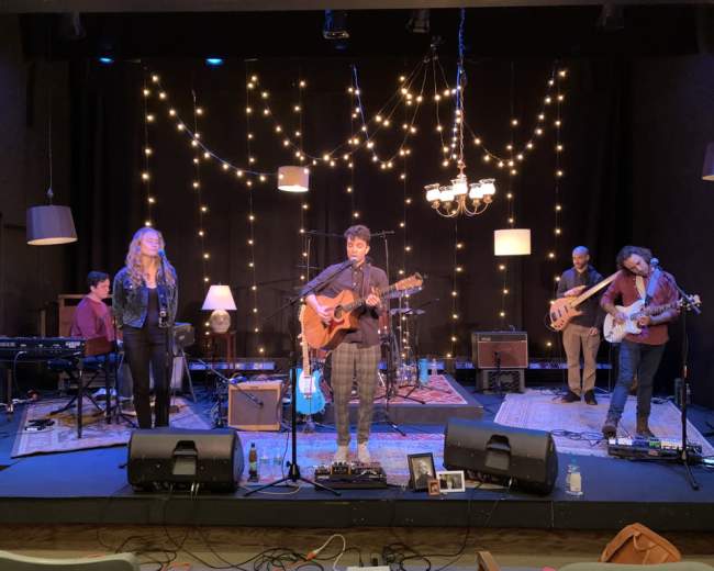 A group of musicians performs on stage with instruments and decorative string lights in the background.
