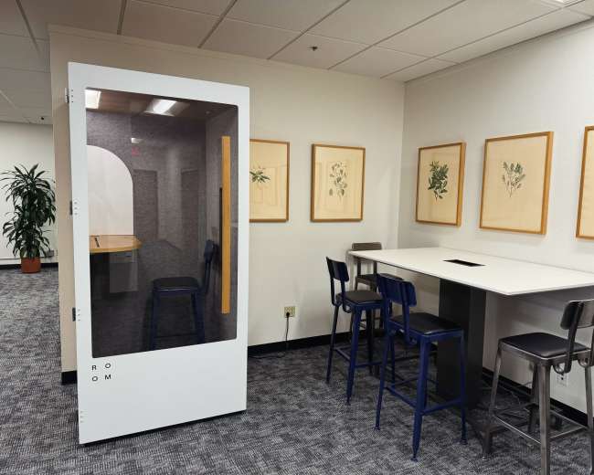 The image shows a small glass-walled meeting room with a round table and chairs beside a larger shared workspace area containing additional seating and framed botanical prints on the walls.