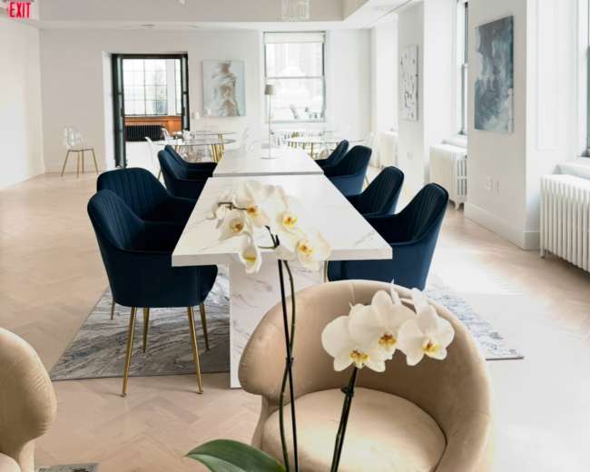 The image shows a modern, light-filled dining or meeting area featuring a long white table, blue chairs, and decorative chandeliers.