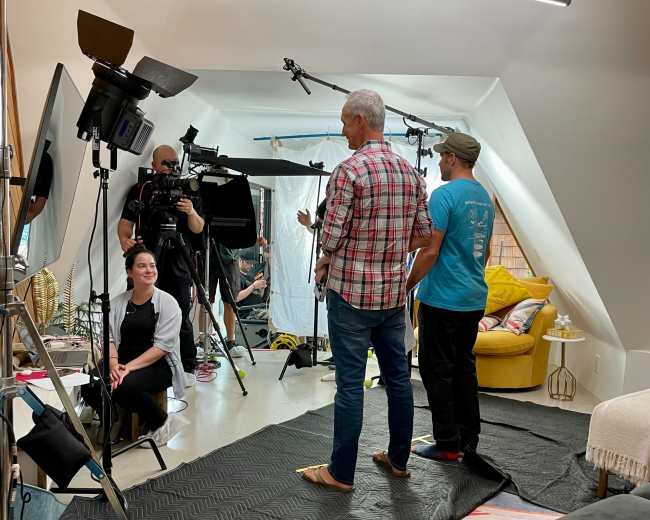 A film crew sets up a scene in a well-lit attic space, featuring two individuals preparing for a shoot while a woman sits nearby.