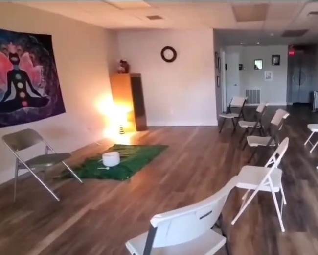 The image shows a spacious room set up with several white folding chairs arranged in a semi-circle around a small green area with a bowl in the center.