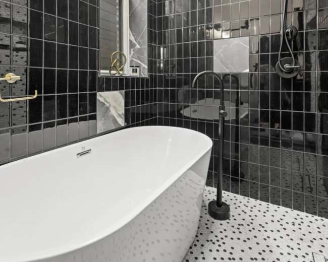 A modern bathroom features a freestanding white bathtub set against black tiled walls with a mosaic floor.