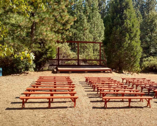 The image shows a clearing with rows of wooden benches facing a small stage surrounded by trees.