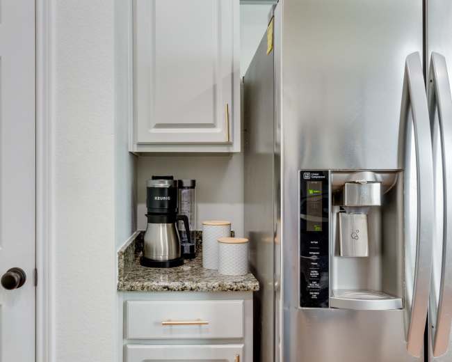 A compact kitchen area with a black coffee maker, two round containers, and a stainless steel refrigerator next to a white cabinet door.