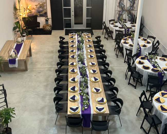 A large dining area features two long tables set with white tablecloths and purple accents, arranged for a formal event in a spacious room with exposed brick walls.