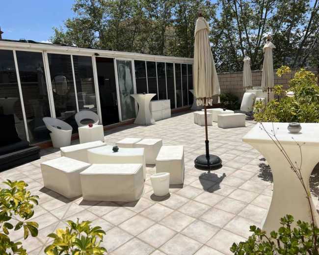 The image shows a sunlit outdoor patio featuring white furniture, including tables and chairs, surrounded by greenery and several sun umbrellas.