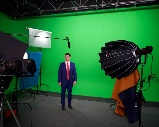 A man in a blue suit and red tie stands in front of a green screen in a photography studio, surrounded by lighting and equipment.