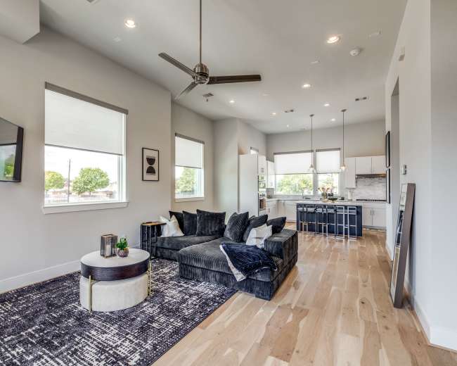 A modern living room features a dark sectional sofa, a round coffee table, and a television mounted on the wall, adjacent to an open kitchen with white cabinets and large windows.