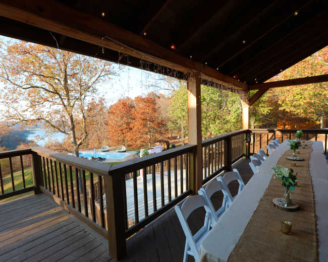 The image shows a wooden deck with a long table set for dinner, overlooking a scenic view of autumn trees and a body of water.