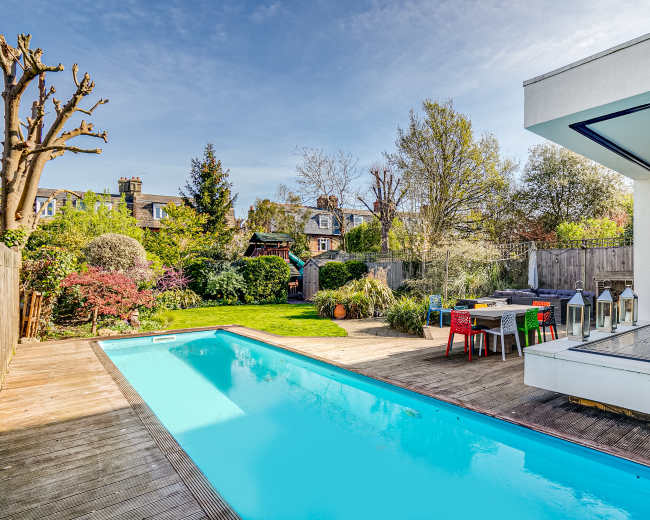 A modern home features a rectangular swimming pool surrounded by a wooden deck and a lush garden with diverse greenery and seating areas.
