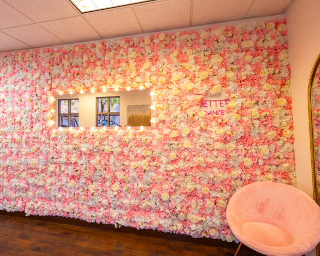 The image features a brightly lit room with a wall covered in artificial pink and white flowers, alongside a circular chair and a window-like frame.