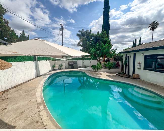 The image shows a residential backyard featuring a clear swimming pool surrounded by concrete decking, tropical plants, and a shaded patio area.
