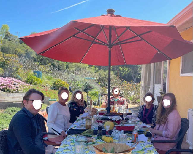 A group of six people sits around a table set with food and drinks under a large red umbrella in a sunny outdoor setting.
