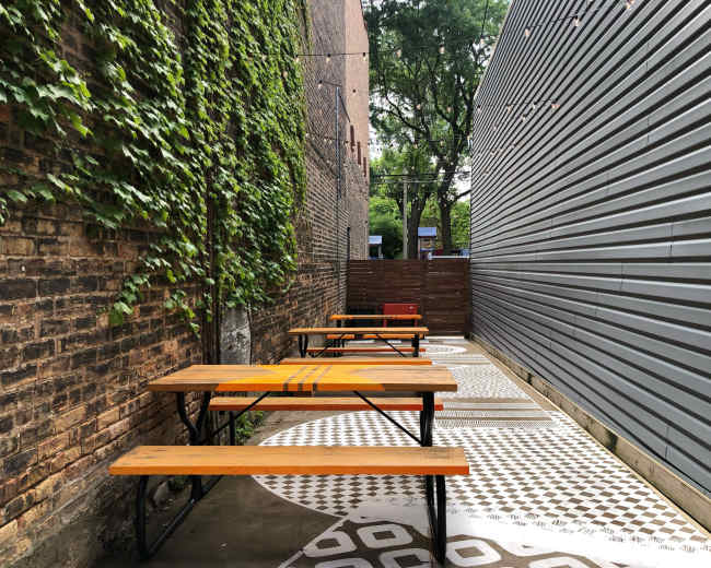 The image shows an outdoor seating area with wooden tables and patterned concrete flooring, enclosed by brick and a metal wall, and flanked by green vines.