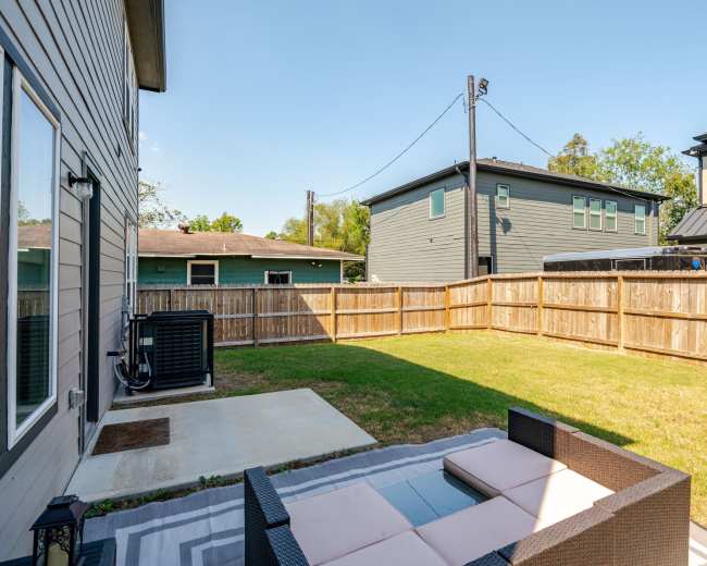 The image shows a backyard with a fenced area featuring a small patio set and grass, surrounded by two houses.