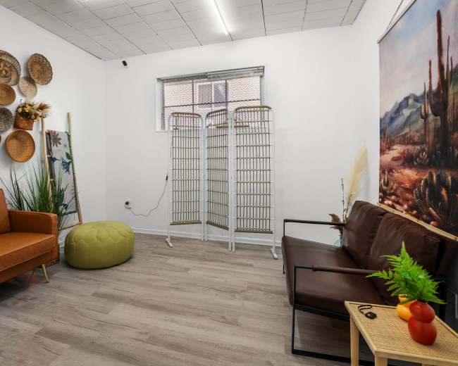 The image shows a small, decorated room featuring a brown couch, an orange armchair, a green pouf, and a wall adorned with woven baskets and a nature-themed painting.