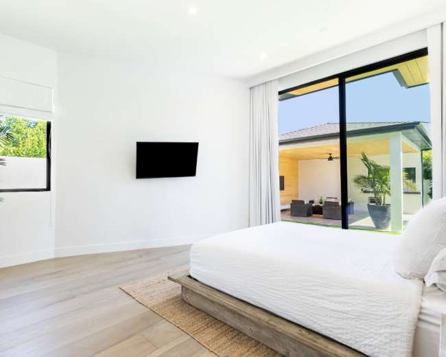 A modern bedroom features large windows with a view of an outdoor seating area, a flat-screen TV mounted on the wall, and a bed with a wooden frame.