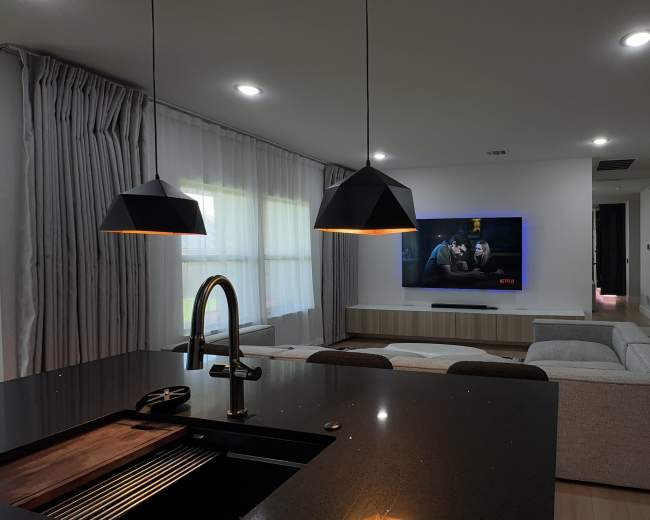 The image shows a modern living room with a gray sofa facing a wall-mounted television, a kitchen area in the foreground featuring a black countertop and pendant lights, and large windows with sheer curtains.