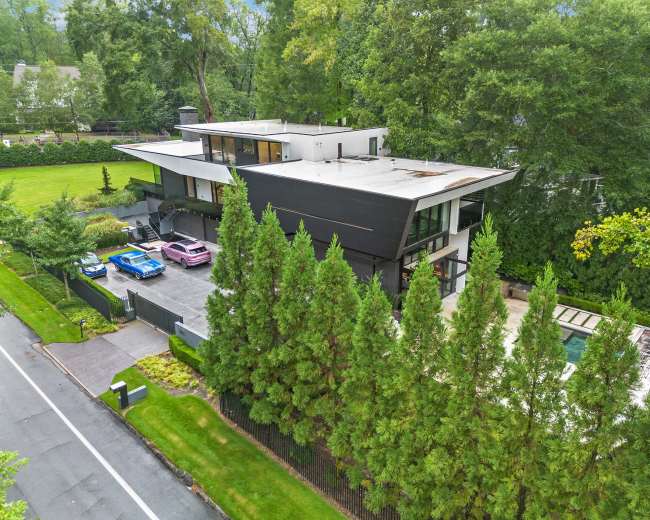The image shows a modern, multi-level house surrounded by trees, with several cars parked in a driveway and a pool visible in the backyard.