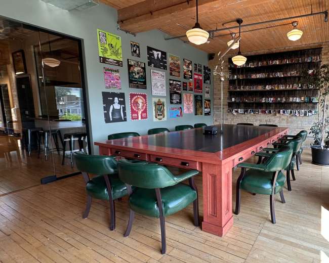 The image shows a spacious dining area featuring a large wooden table surrounded by green chairs, with brick walls adorned with posters and a wall of shelves in the background.