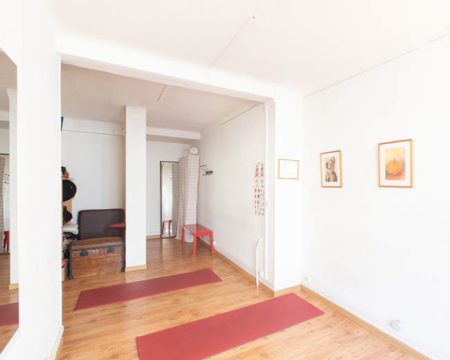 The image shows a bright yoga studio with wooden flooring, red yoga mats laid out, and several framed artworks on the walls.