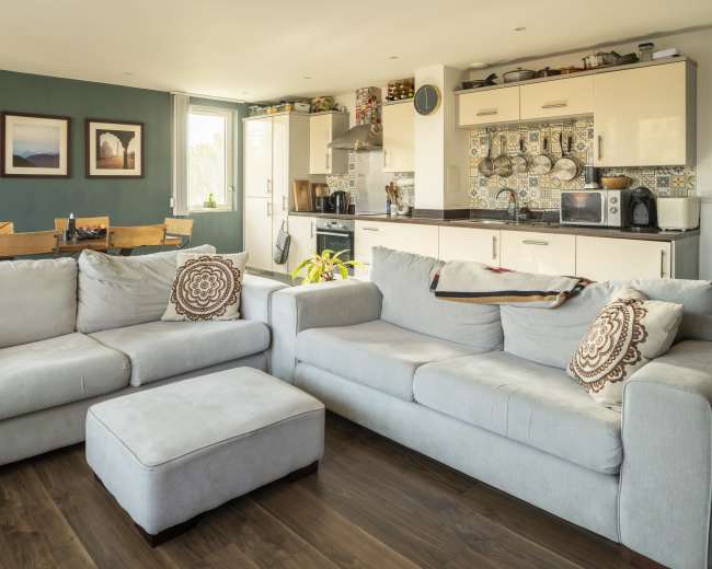 The image shows a modern living room with two light-colored sofas, a coffee table, and an adjoining kitchen area featuring cabinets and a dining table.