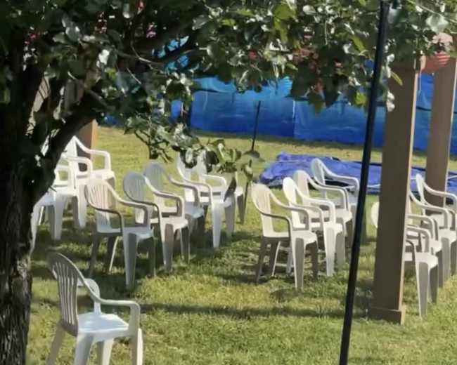 A grassy area with several white plastic chairs arranged under a tree, and a blue tarp in the background.