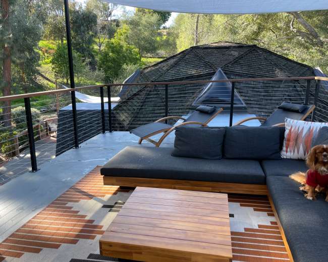 A wooden deck features a sectional sofa, a coffee table, and two lounge chairs overlooking a wooded area and a distinctive roof structure.