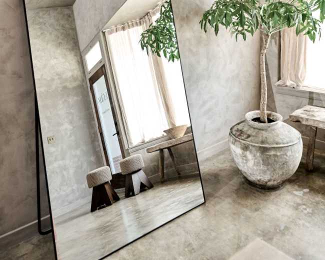 A large, arched mirror leans against a wall in a room with minimalistic decor, featuring a potted plant, a wooden table, and natural light streaming through sheer curtains.