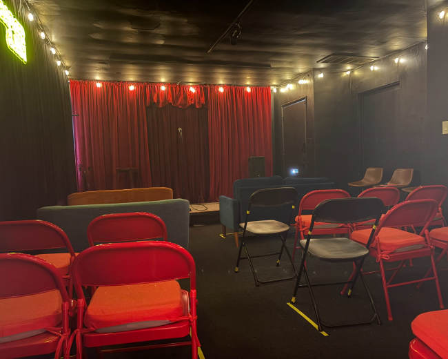 The image shows a small performance space with a stage, red curtains, and seating arranged in rows, including red and black chairs, along with a few sofas at the back.