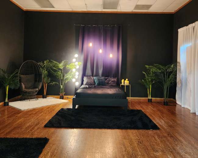 A dark-themed bedroom with a black bed, hanging lights, and various indoor plants, complemented by a hanging chair and rugs on the wooden floor.