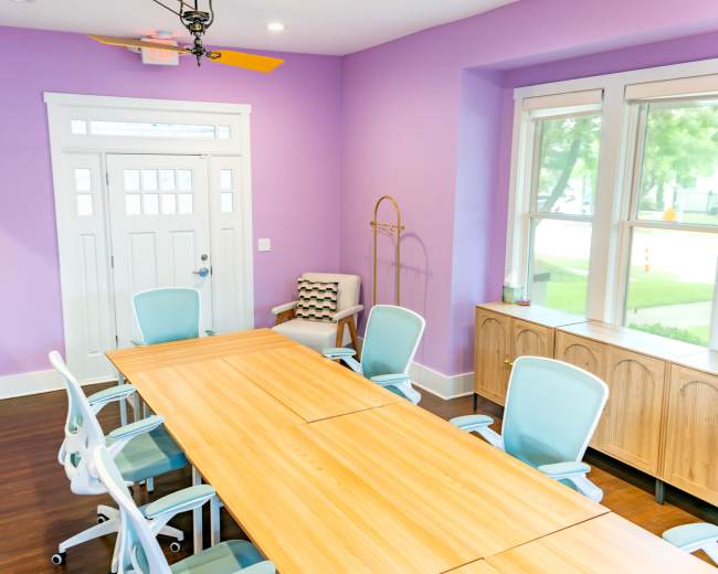 A conference room features a large wooden table surrounded by light blue chairs, with purple walls and a white door leading outside.